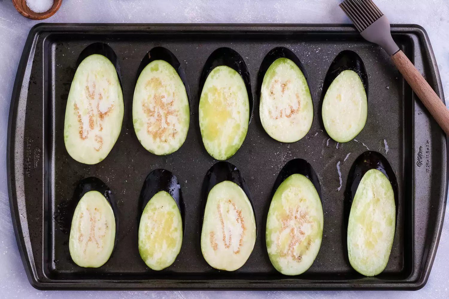 Fatias de berinjela em uma assadeira pinceladas com azeite e polvilhadas com sal