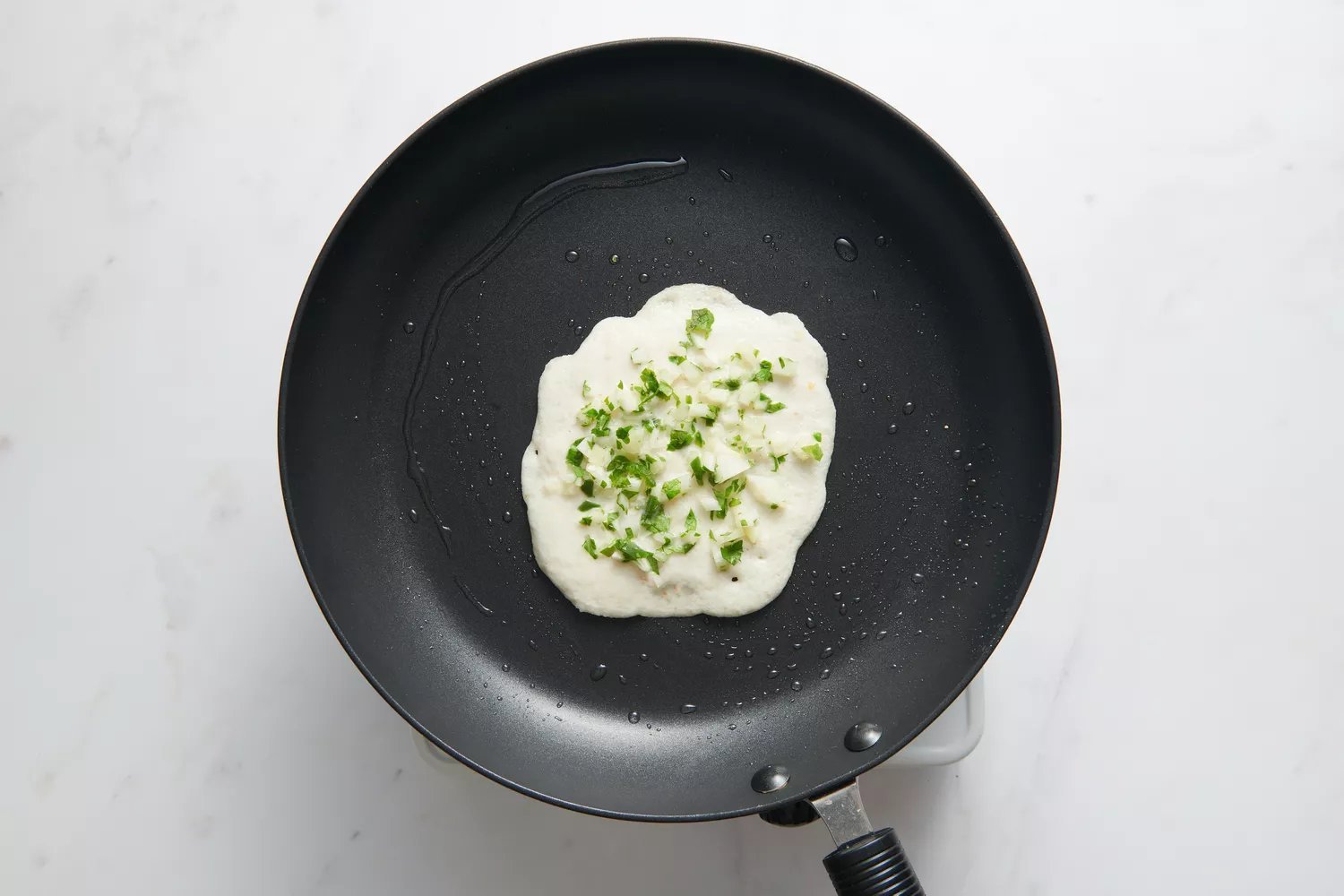 Uma frigideira antiaderente com uma pequena porção de massa no centro, coberta com cebolas picadas, pimentas e coentro